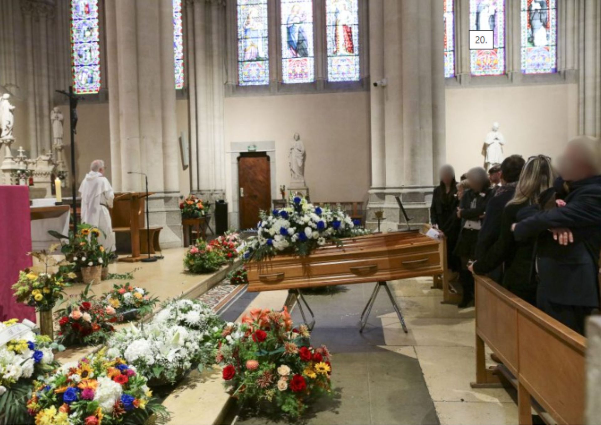 Cérémonie funéraire à l'église avec cercueil, compositions florales, personnes en deuil et prêtre.