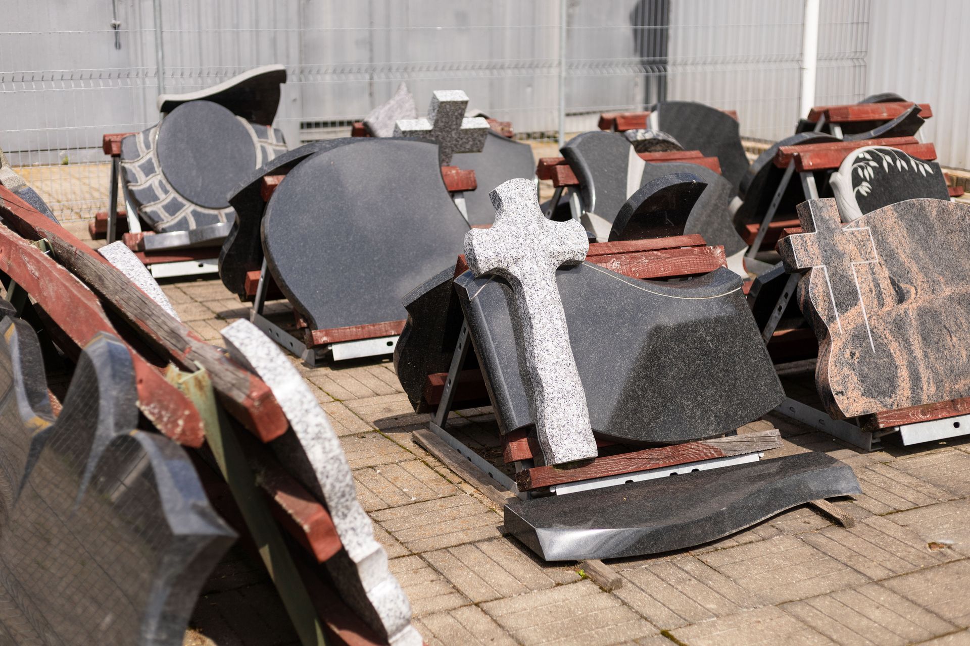 Des pierres tombales de formes et de couleurs variées, entreposées à l'extérieur sur des supports métalliques.