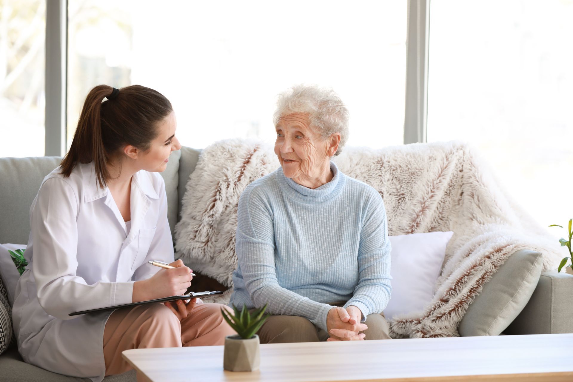 Une femme en blouse blanche consulte une femme plus âgée assise sur un canapé ; intérieur.