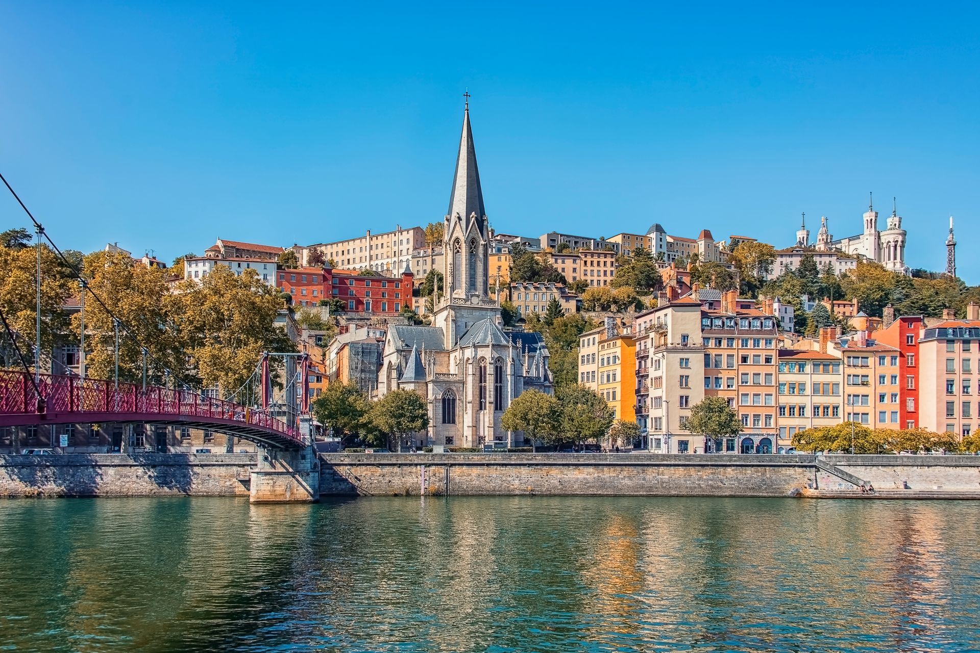 Paysage urbain de Lyon, en France, avec une église à la flèche élancée, des bâtiments colorés le long d'une rivière et un pont.