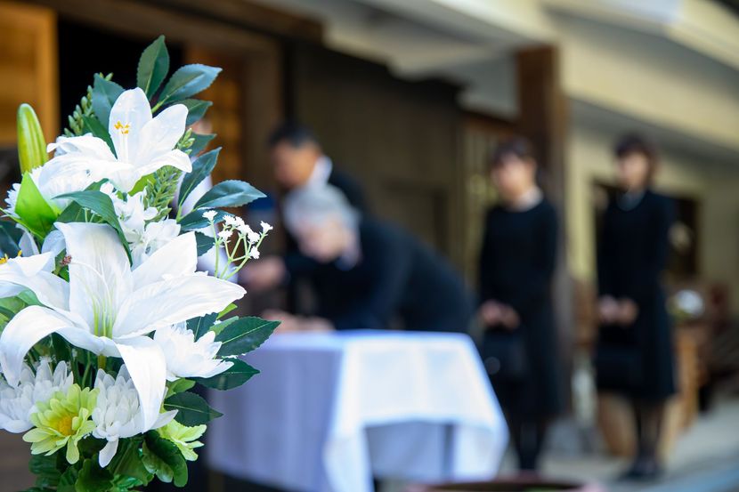 Des lys blancs au premier plan ; des personnes vêtues de noir se rassemblent près d'une table à l'extérieur d'un bâtiment.