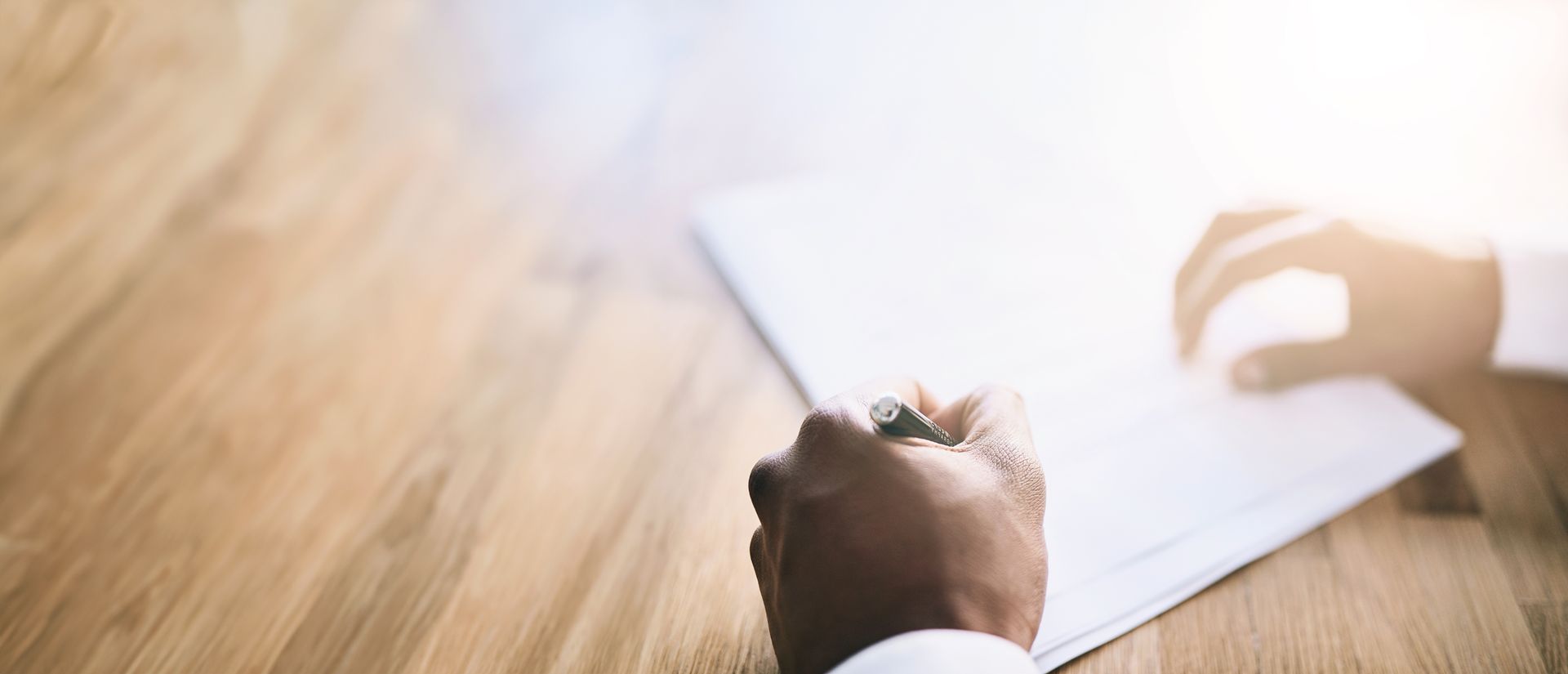 Écriture manuscrite sur une feuille de papier blanc vierge, posée sur une surface en bois, avec un stylo, à la lumière du soleil.