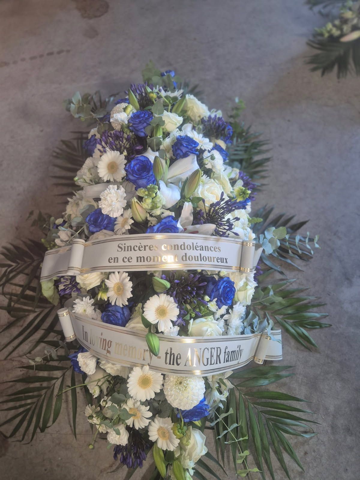 Composition florale funéraire avec des fleurs blanches et bleues et une bannière.