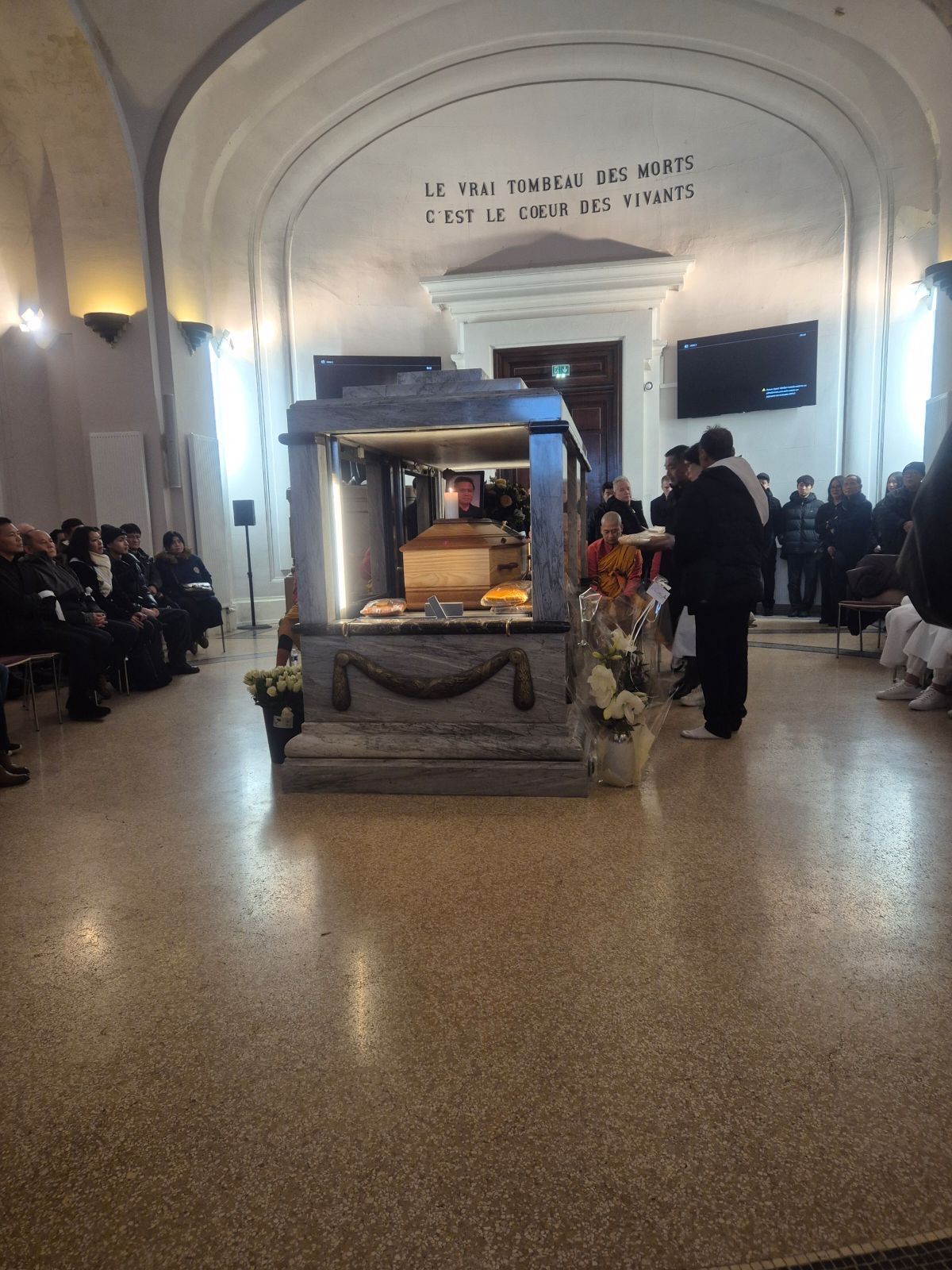 Cérémonie avec un cercueil sur une estrade, personnes assises et debout dans une salle voûtée.