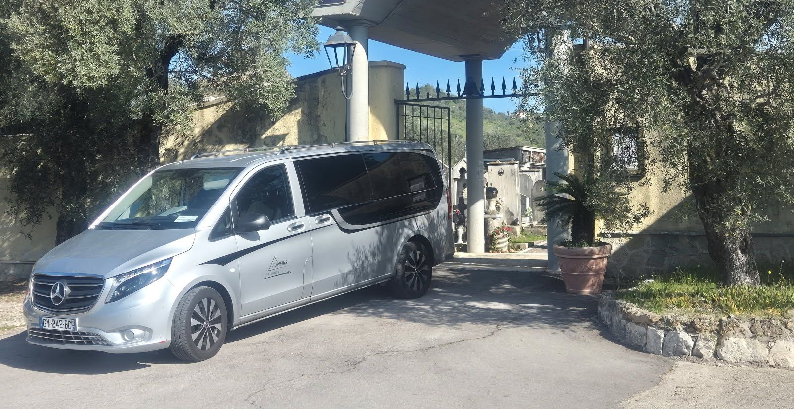 Une camionnette argentée est garée devant une entrée fermée par un portail, des arbres de chaque côté.