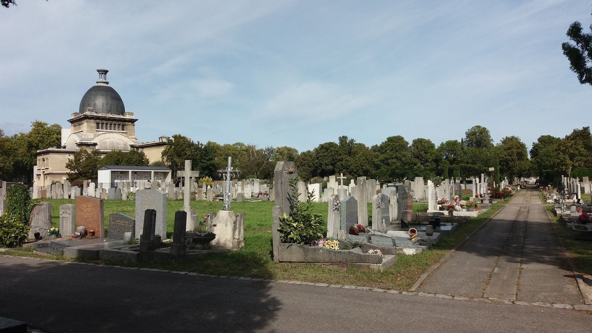 Scène de cimetière avec de nombreuses pierres tombales et un bâtiment à dôme en arrière-plan, sous un ciel bleu.