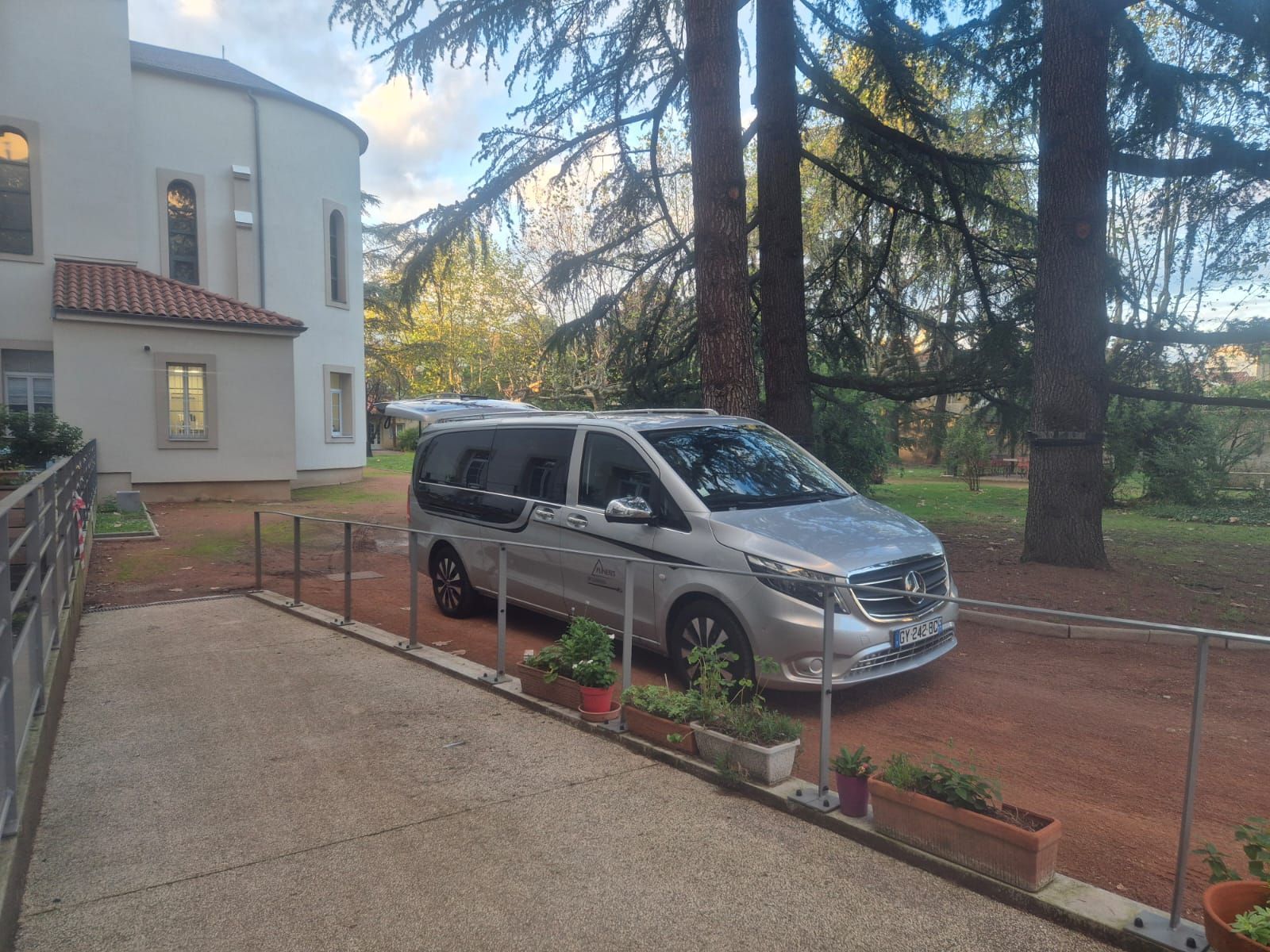 Une minifourgonnette argentée est garée dans une allée devant un bâtiment blanc, avec des arbres en arrière-plan.