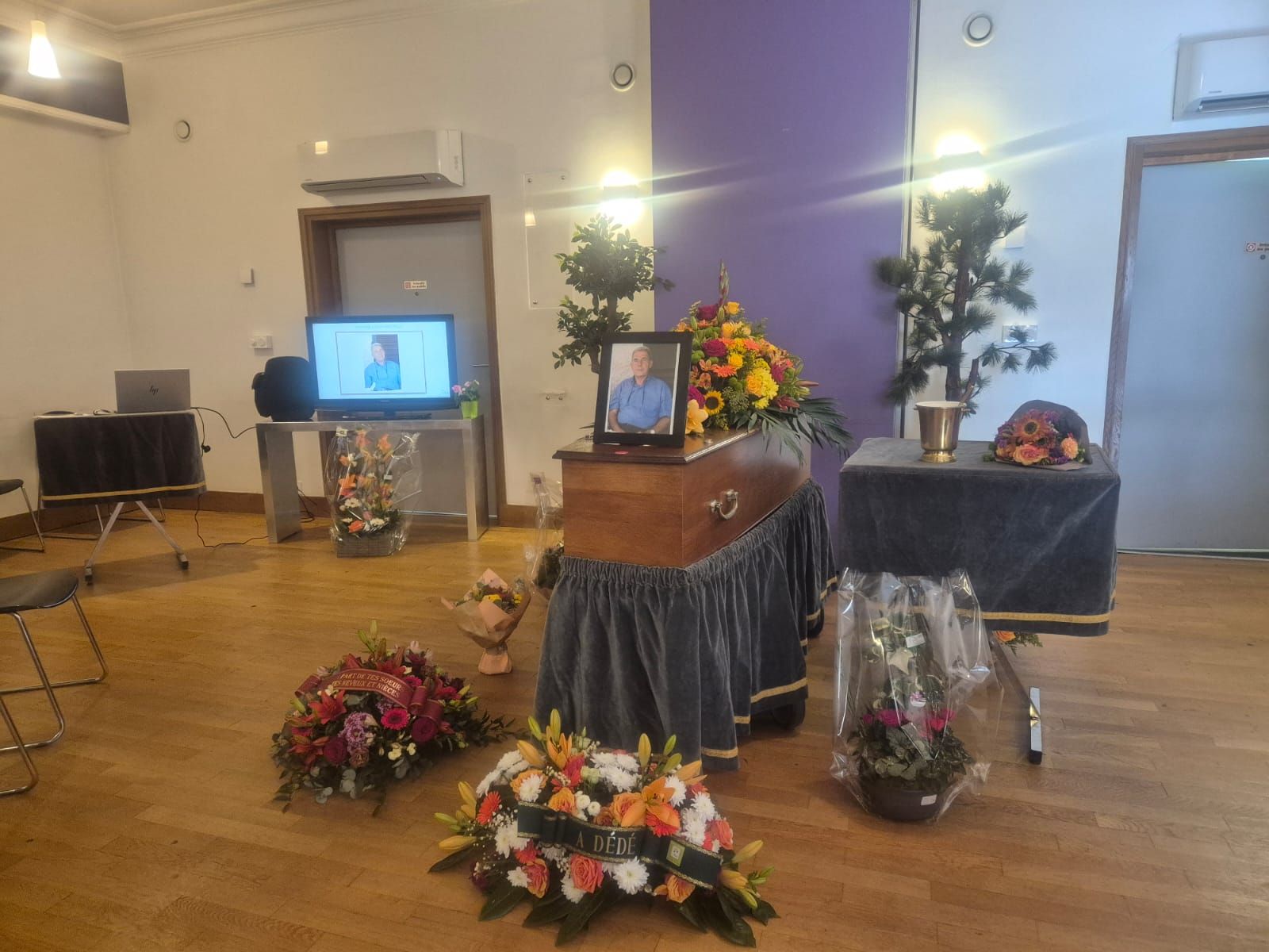 Une cérémonie funéraire : un cercueil fermé avec des compositions florales. Une photo et un écran.