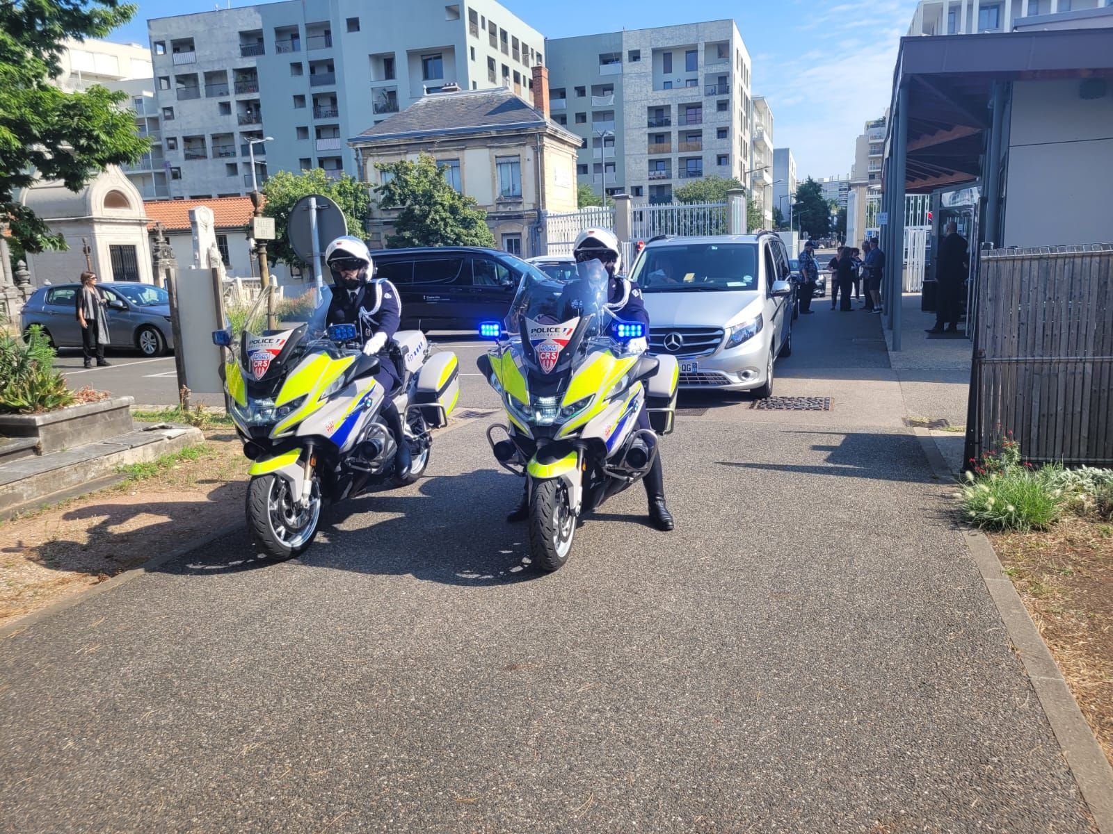 Deux motos de police, gyrophares allumés, sont stationnées près d'une camionnette. Un bâtiment se dresse en arrière-plan.