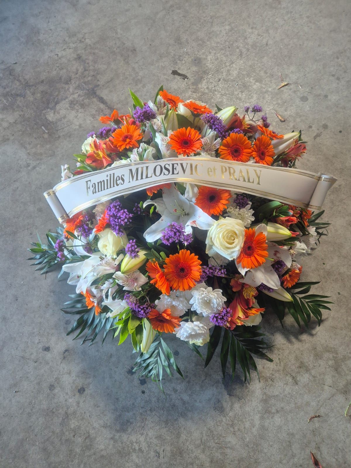 Couronne de fleurs avec des fleurs orange, blanches et violettes ; le ruban blanc porte l'inscription « Familles Milosevic de Praly ».