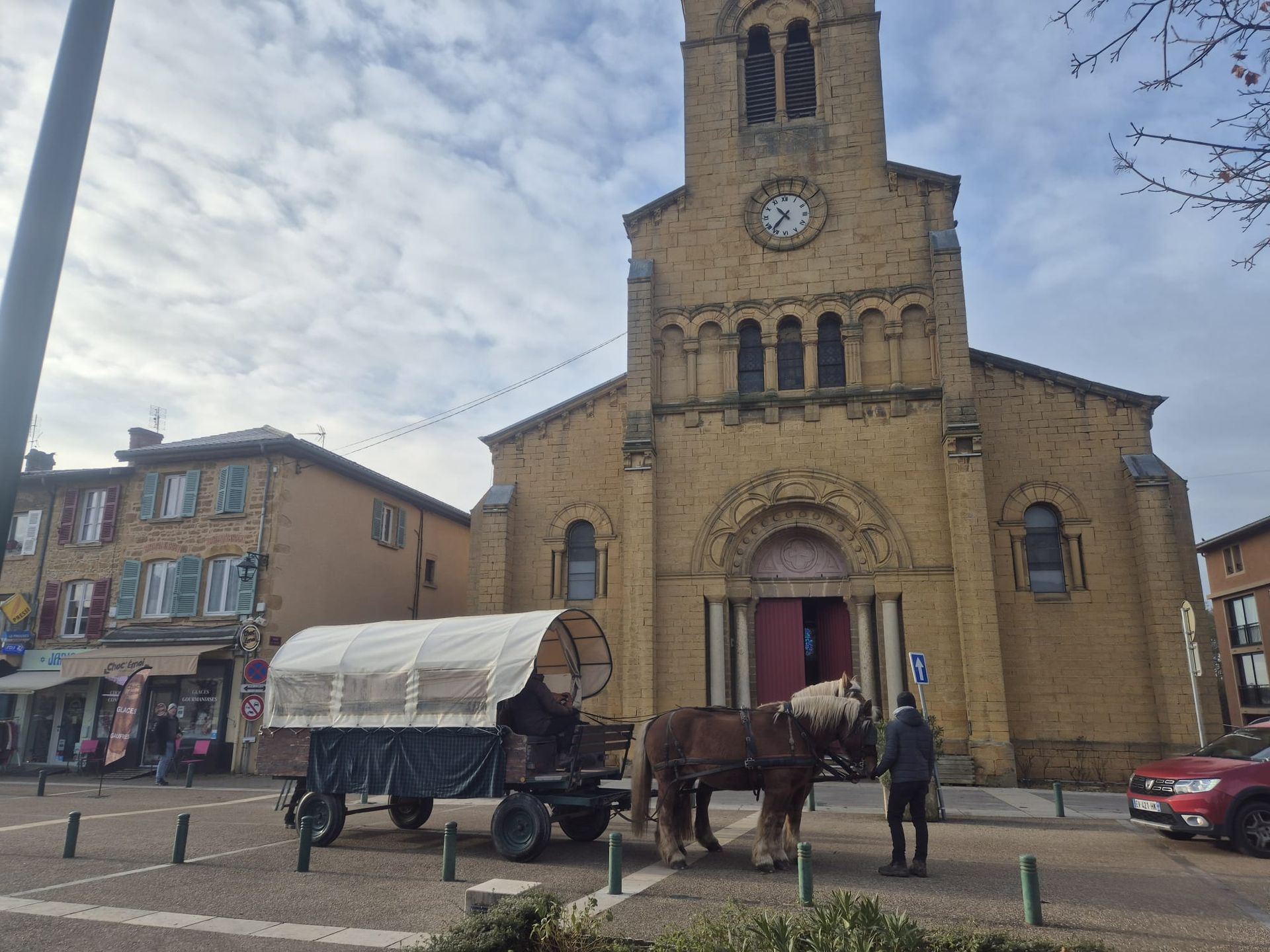 Calèche devant une église ; une personne se tient à proximité. Bâtiments et ciel nuageux en arrière-plan.
