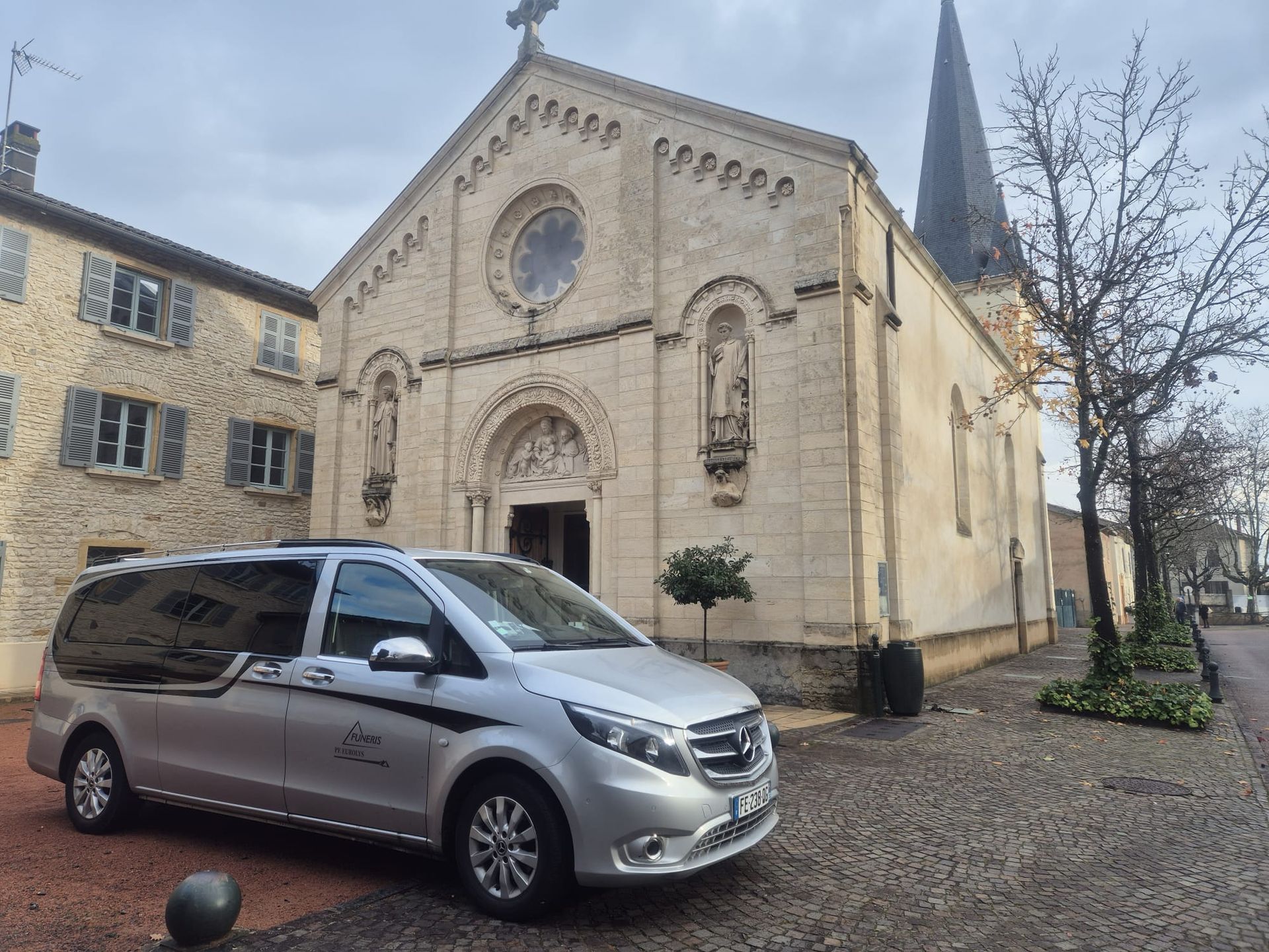 Un corbillard argenté est garé devant une église claire à clocher, située dans une rue pavée.