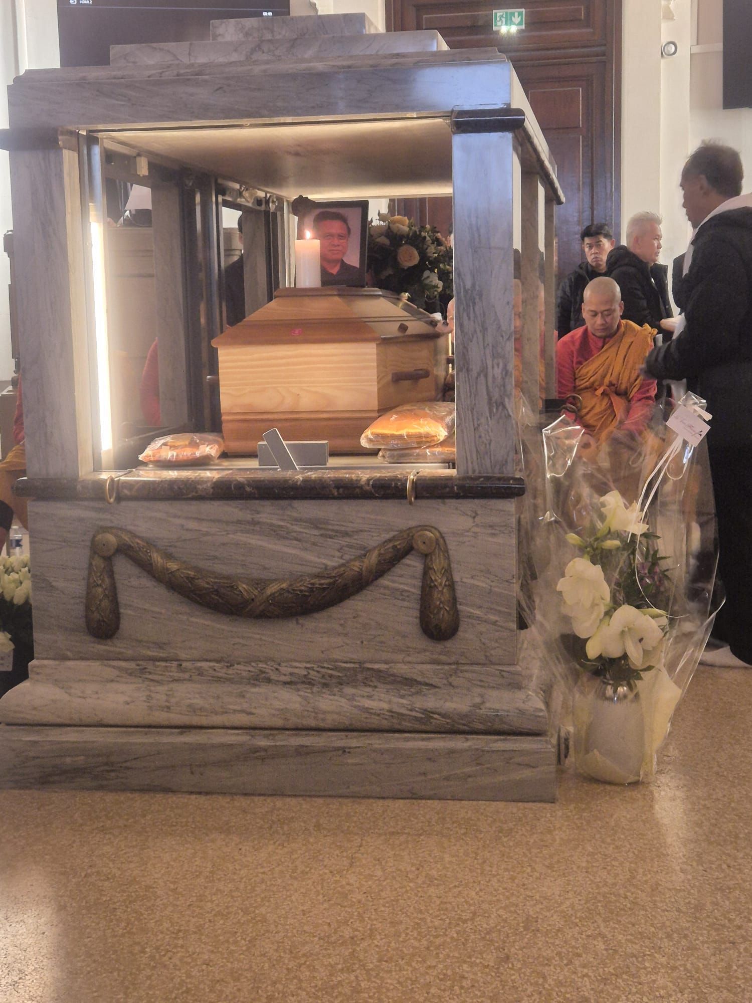 Cercueil en bois exposé dans une structure en marbre, fleurs et personnes en deuil dans un cadre religieux.
