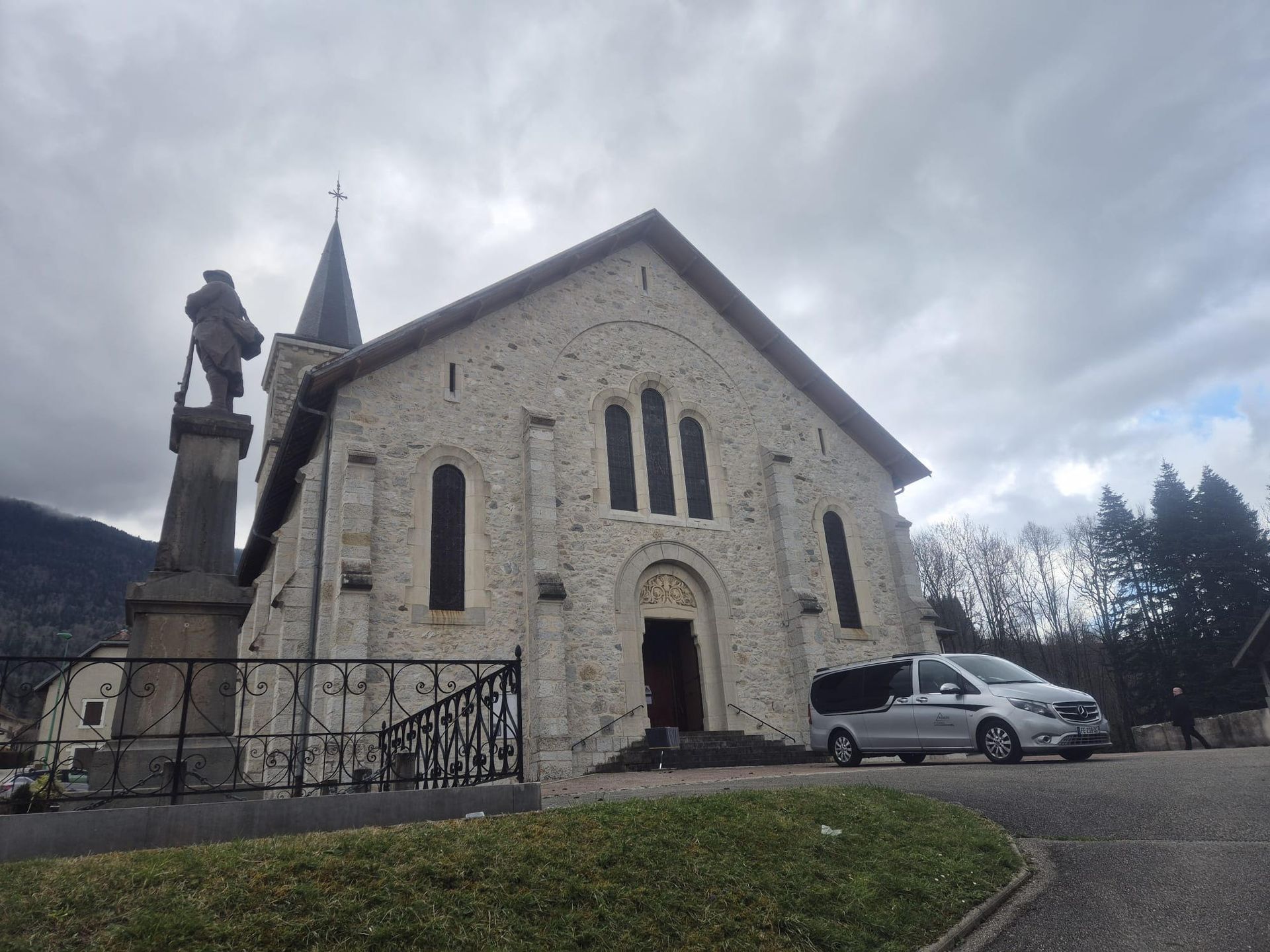 Église en pierre avec une flèche, une statue et une camionnette garée devant par temps nuageux.