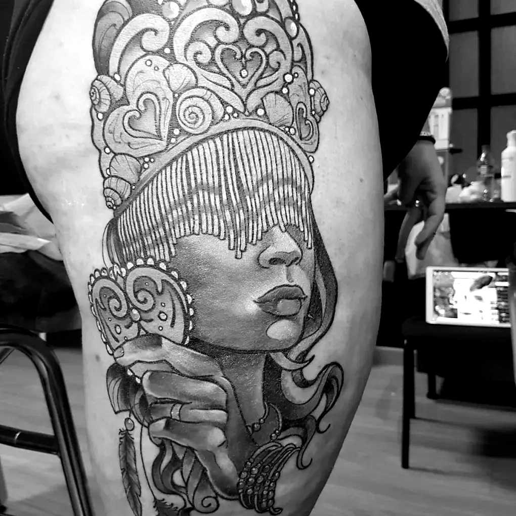 Un tatuaje en blanco y negro en el muslo que representa a una persona con una corona ornamentada.