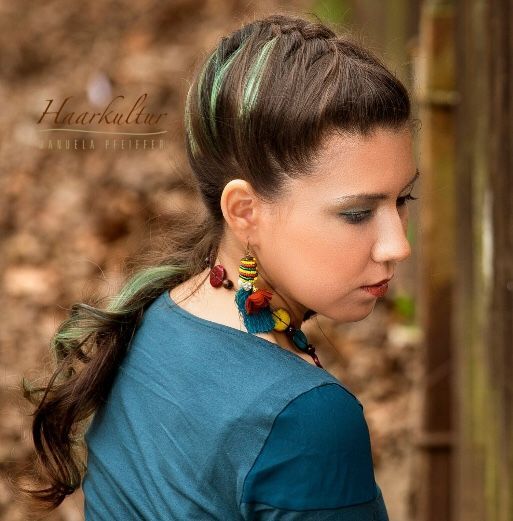 Frau mit türkisfarbenem Kleid, bunten Ohrringen und teilweise geflochtenem braunem Haar mit grünen Strähnen, im Freien.