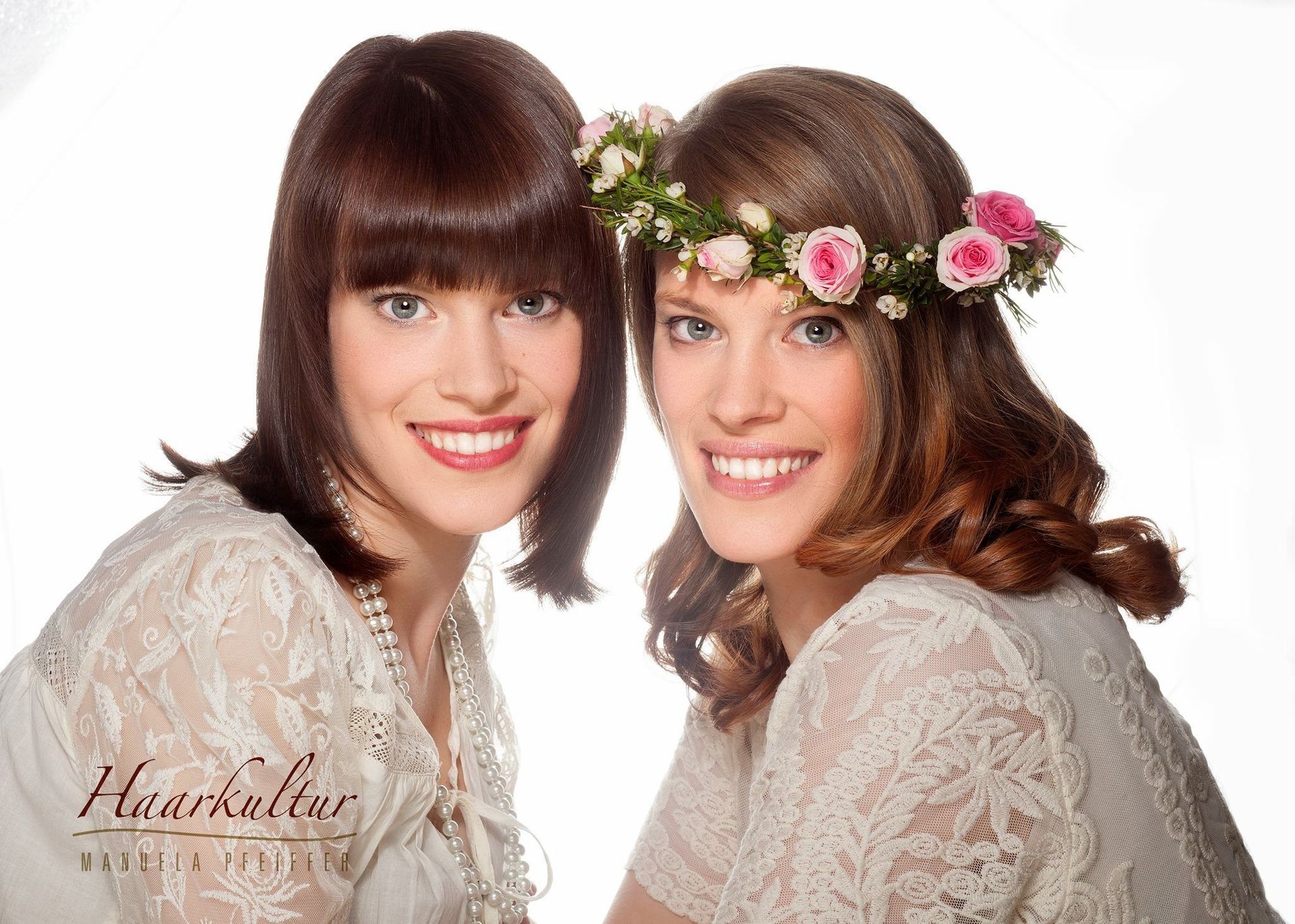 Zwei lächelnde Frauen, eine mit einem Blumenkranz, beide in weißen Spitzenoberteilen, vor weißem Hintergrund.