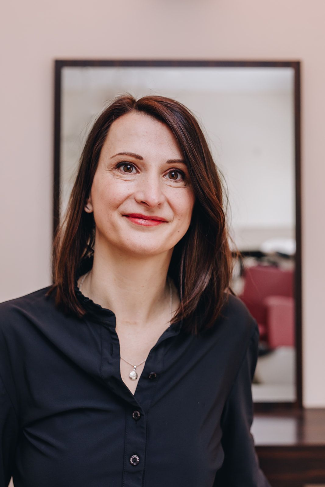 Eine Frau mit braunen Haaren lächelt in einem schwarzen Hemd vor einem Spiegel.