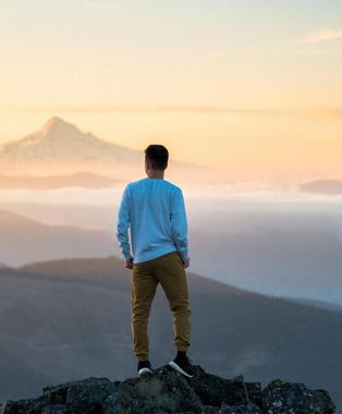 Ein Mann steht auf einem Felsgipfel und blickt bei Sonnenaufgang auf eine Bergkette.