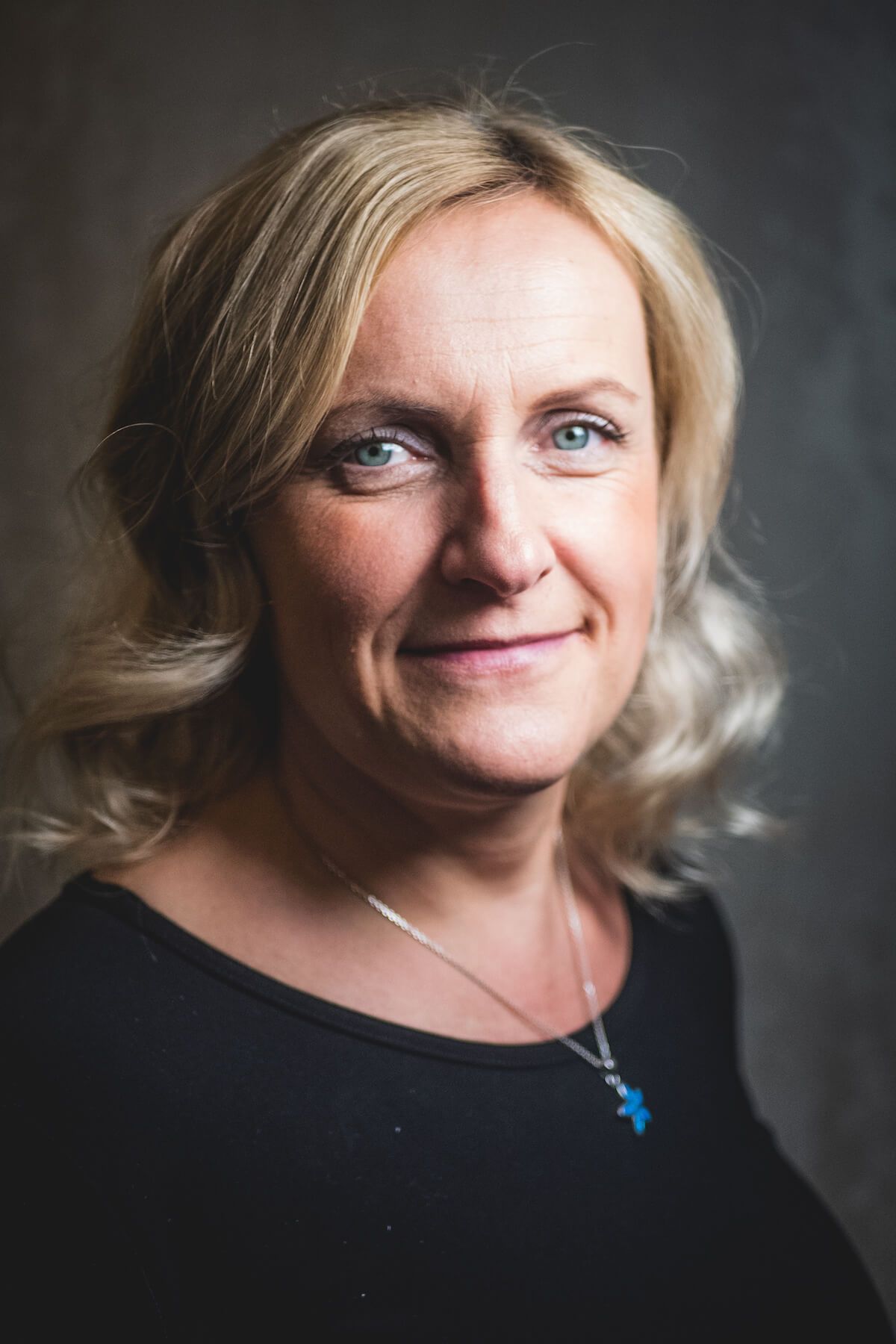 Blonde Frau mit blauen Augen lächelt, trägt ein schwarzes Oberteil und einen Anhänger. Weicher Hintergrund.