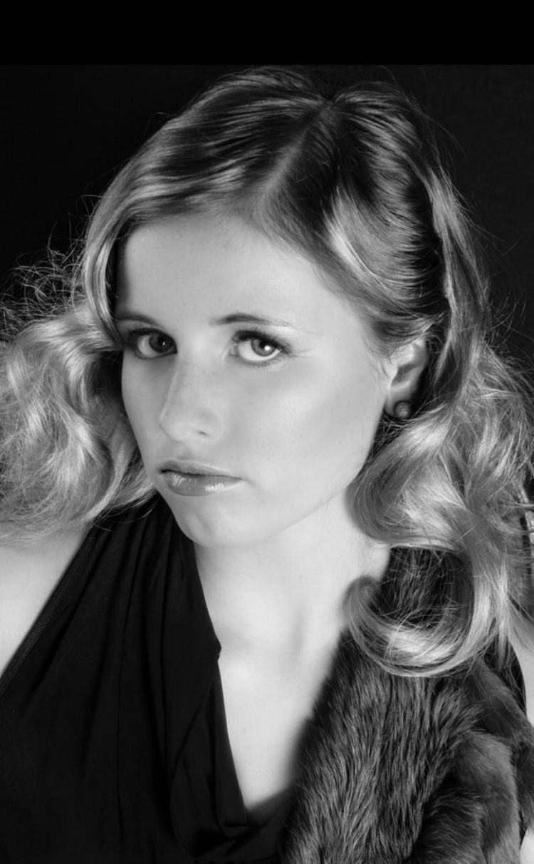 Frau mit welligem blonden Haar, die in die Kamera blickt, trägt ein dunkles Oberteil mit einem Pelzstola.