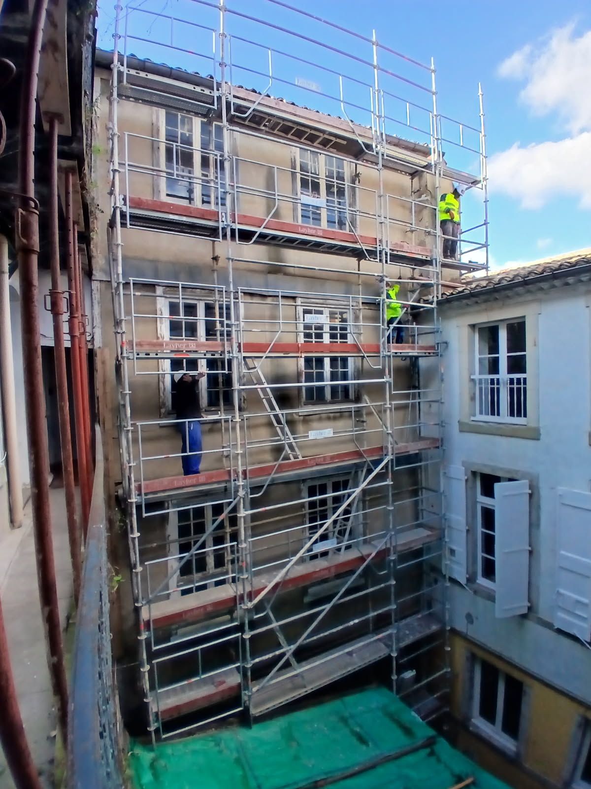 Échafaudage sur la façade d'un bâtiment ; deux ouvriers en gilets haute visibilité. Extérieur avec fenêtres et ciel bleu.