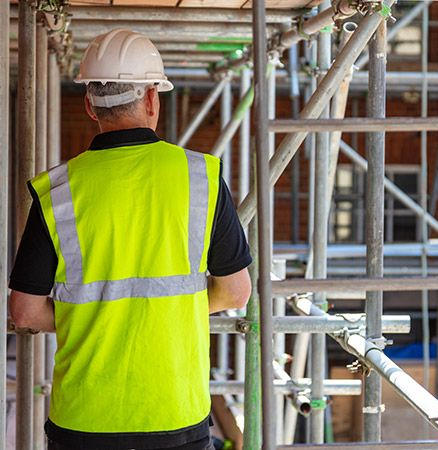 Un homme de dos portant un gilet jaune et un casque de chantier blanc au milieu d'échafaudages