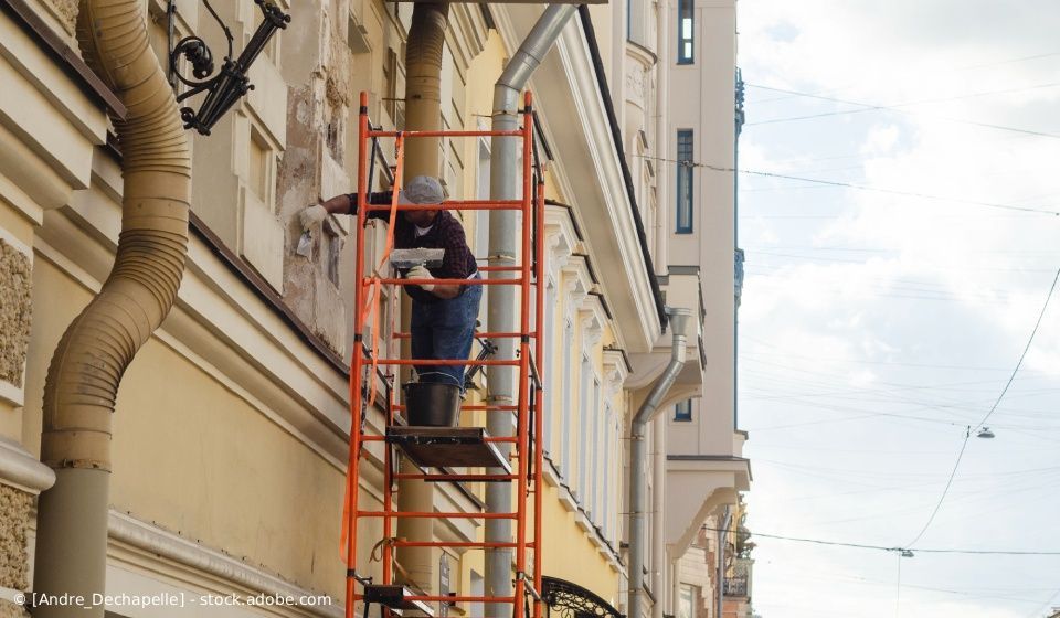 Eine Person repariert auf einem Gerüst die Fassade eines Gebäudes. Orangefarbenes Gerüst