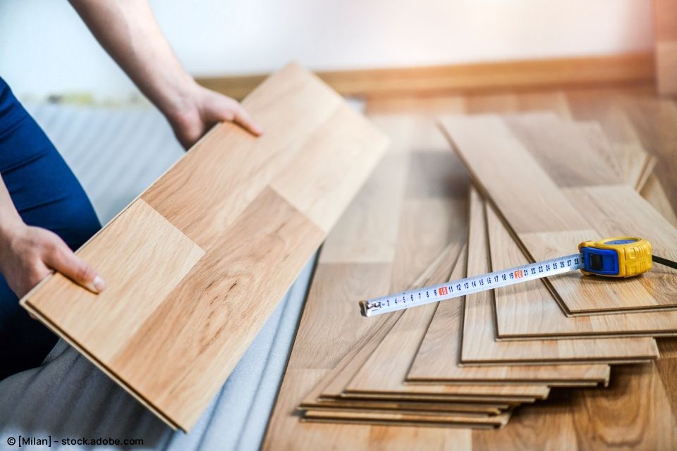 Eine Person hält eine Holzdiele, ein Maßband und Bodenbelagsmaterialien. Innenraum mit hellen Holztönen.