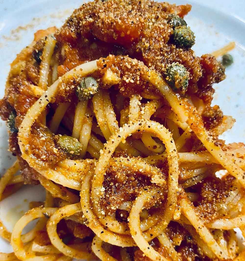 Plato de espaguetis con salsa roja, alcaparras y cobertura de pan rallado.