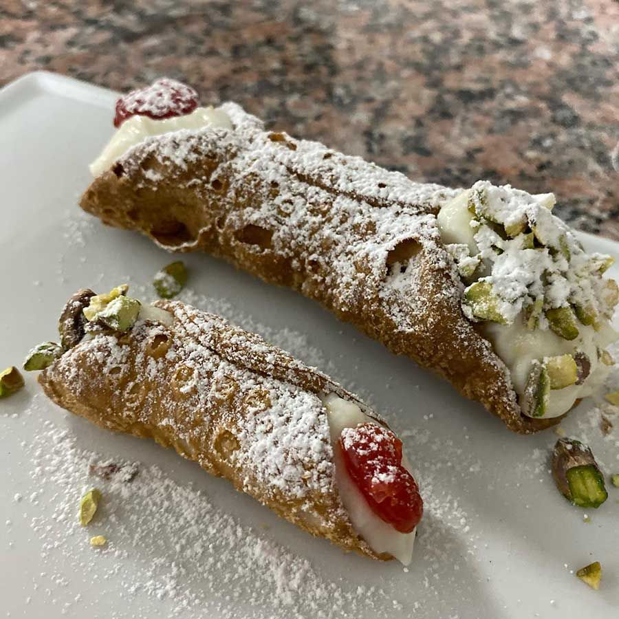 Dos pasteles cannoli, uno grande y otro pequeño, espolvoreados con azúcar en polvo, cubiertos con pistachos y fruta confitada.