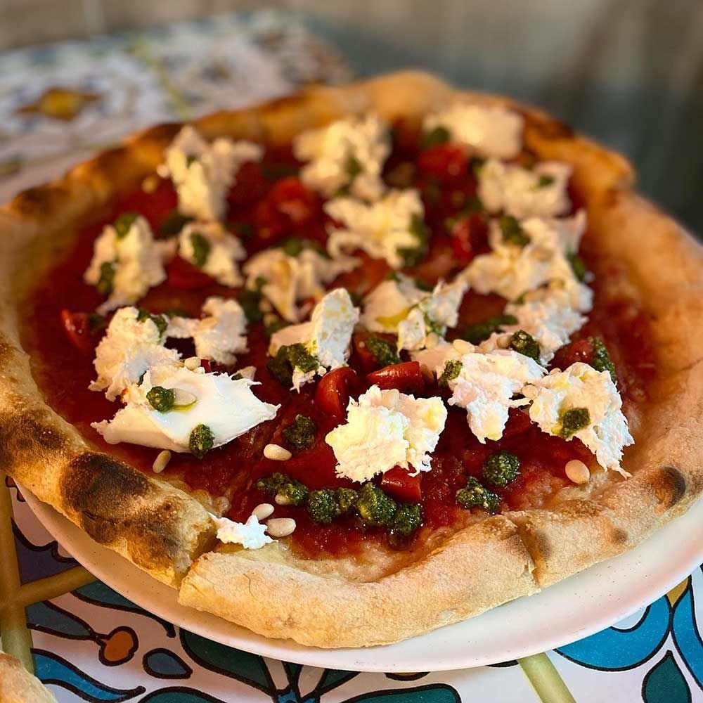 Pizza con salsa roja, queso blanco, pesto y tomates, en un plato blanco, sobre una mesa de azulejos de colores.