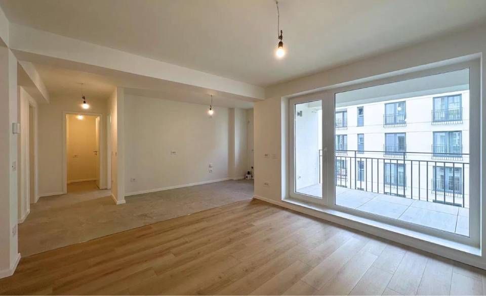 Leeres Zimmer mit Holzboden, grossem Fenster zum Balkon und Zugang zum Flur.