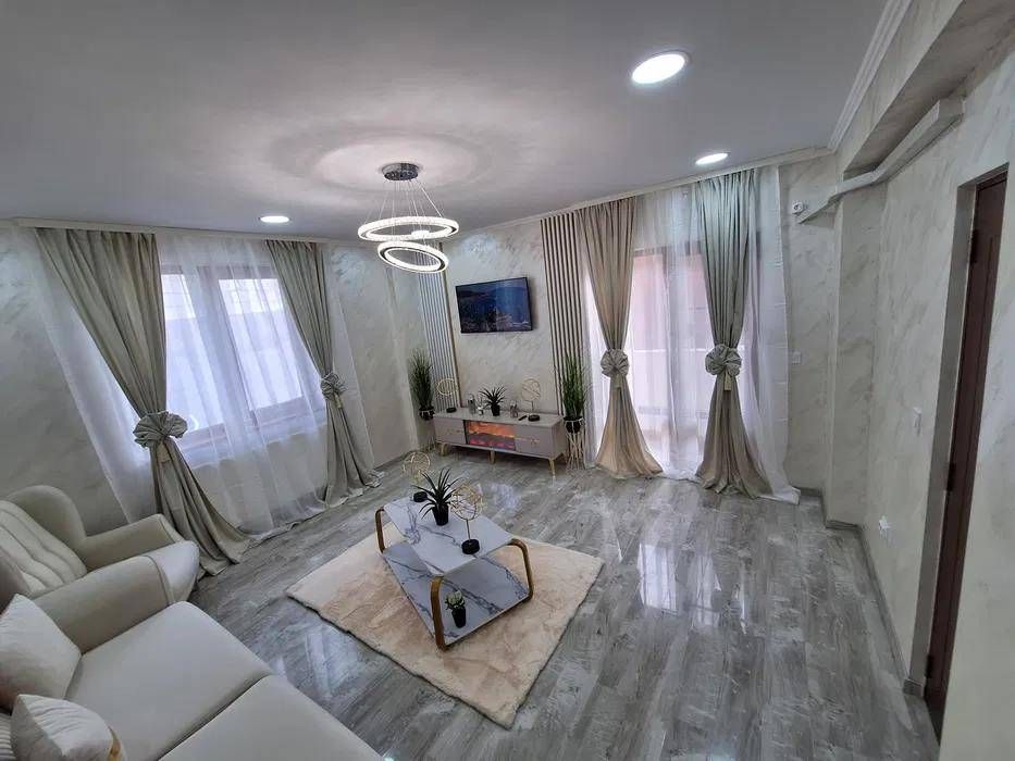 Gemütliches Wohnzimmer mit beigefarbenem Sofa, Marmorboden und hellen Vorhängen. Moderne Einrichtung, Fernseher und Couchtisch.