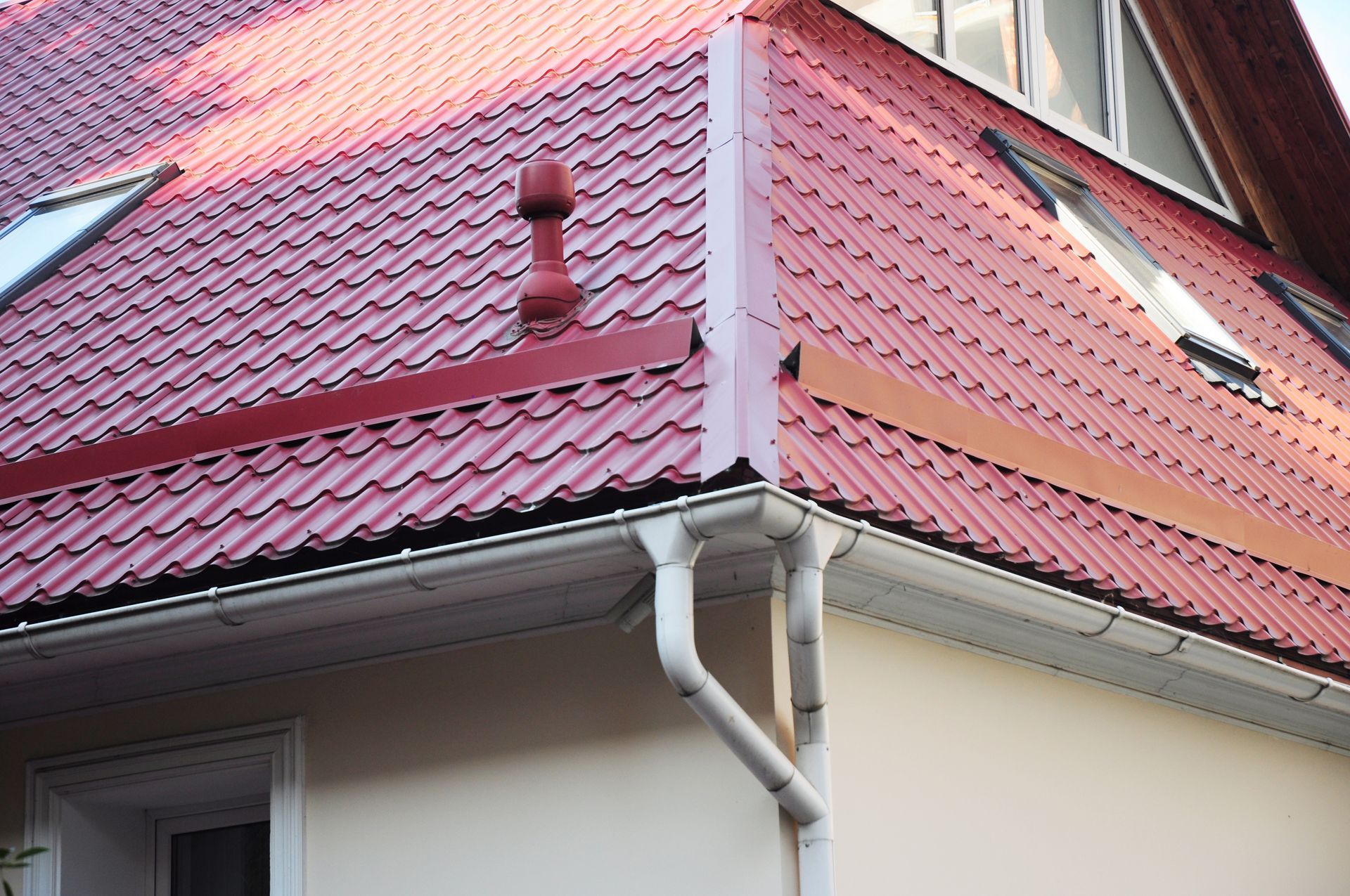 Toit en métal rouge avec gouttières et descente pluviale blanches sur la façade d'une maison.