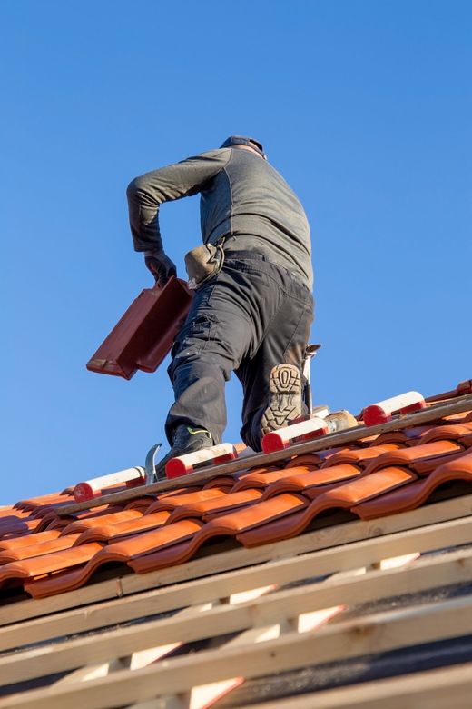 Un ouvrier se tient debout sur un toit de tuiles rouges portant une sacoche à outils rouge.