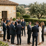 Pequeña reunión formal de personas vestidas de traje conversando al aire libre junto a una villa de piedra y un jardín.
