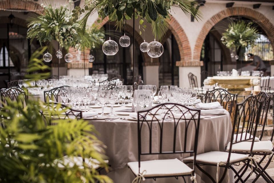 Elegante comedor al aire libre con mesas blancas, sillas negras, faroles de cristal colgantes y arcos de piedra.