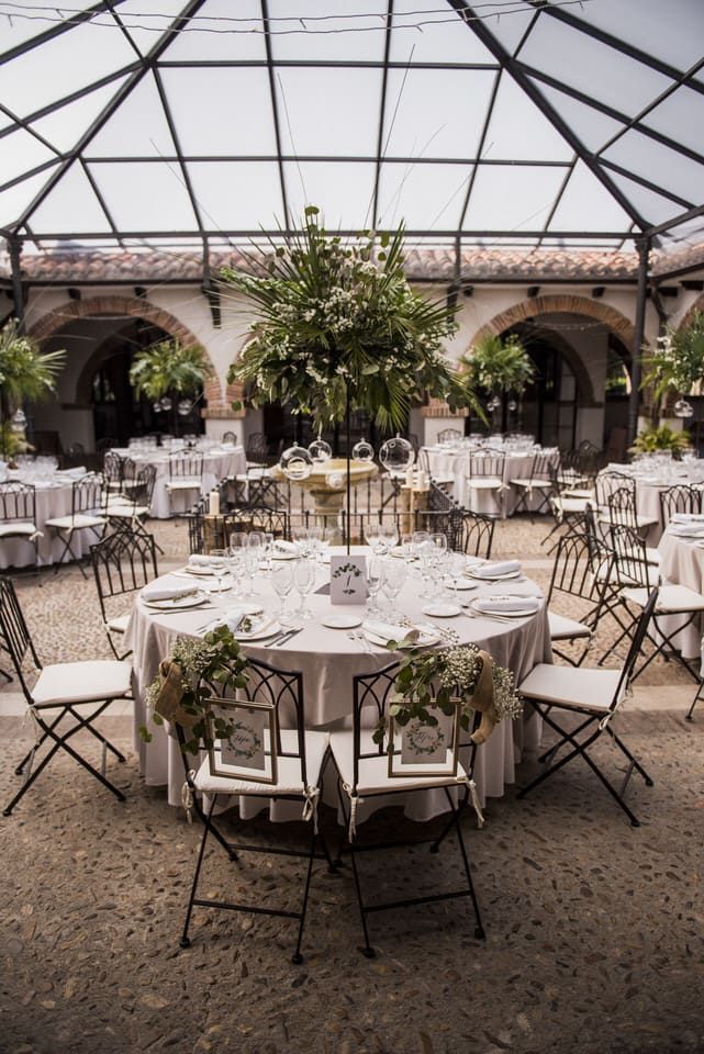 Mesas redondas para banquetes en un patio con techo de cristal, vestidas con manteles blancos y adornadas con arreglos florales y follaje.
