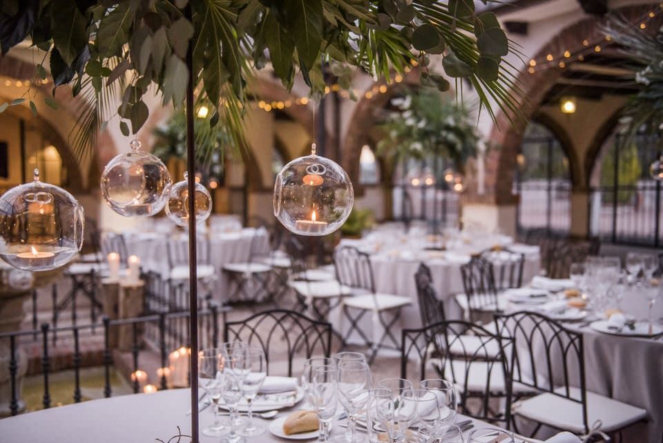 Elegante comedor al aire libre con mesas blancas, lámparas esféricas colgantes y abundante vegetación al atardecer