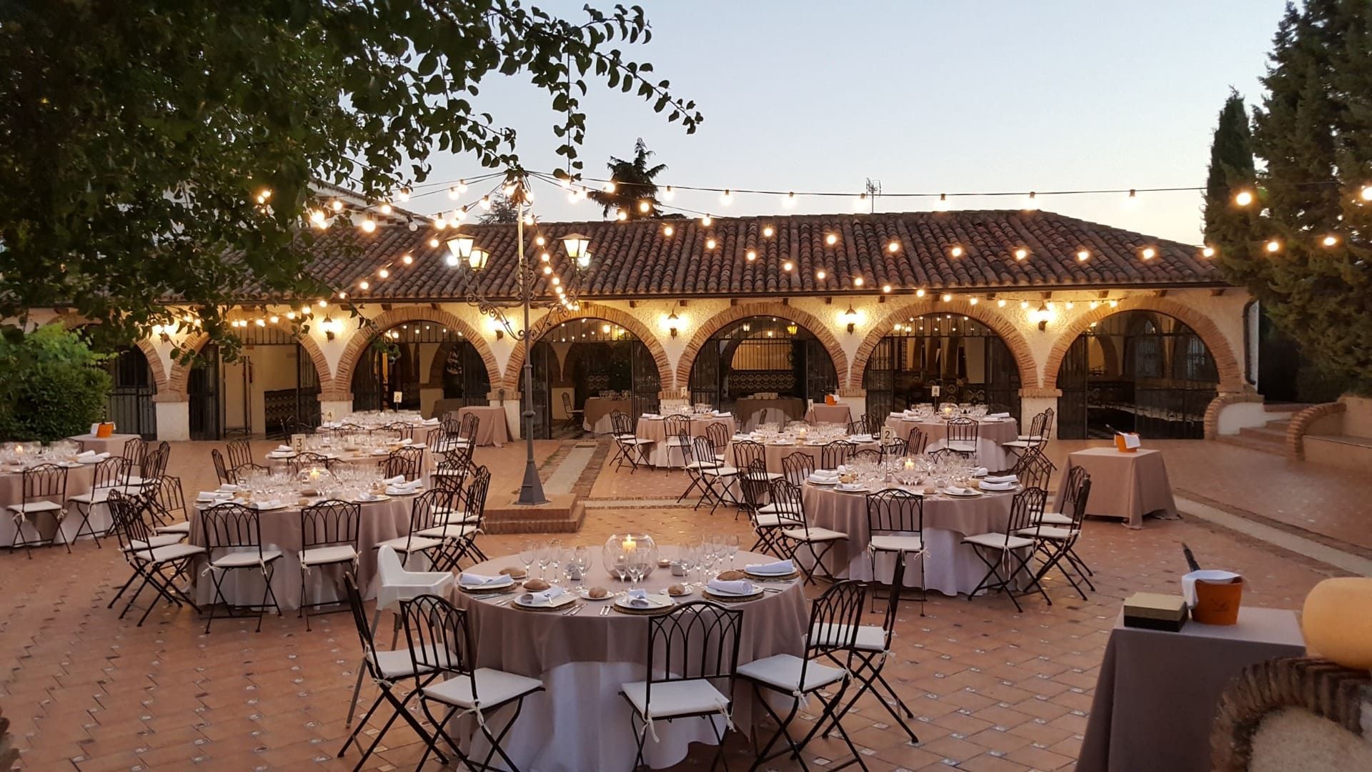 Recepción de boda al aire libre, con mesas redondas, luces de verbena y un lugar rústico al anochecer