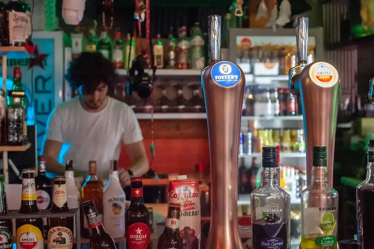 Un camarero con una camiseta blanca está de pie detrás de la barra, con varias botellas de licor