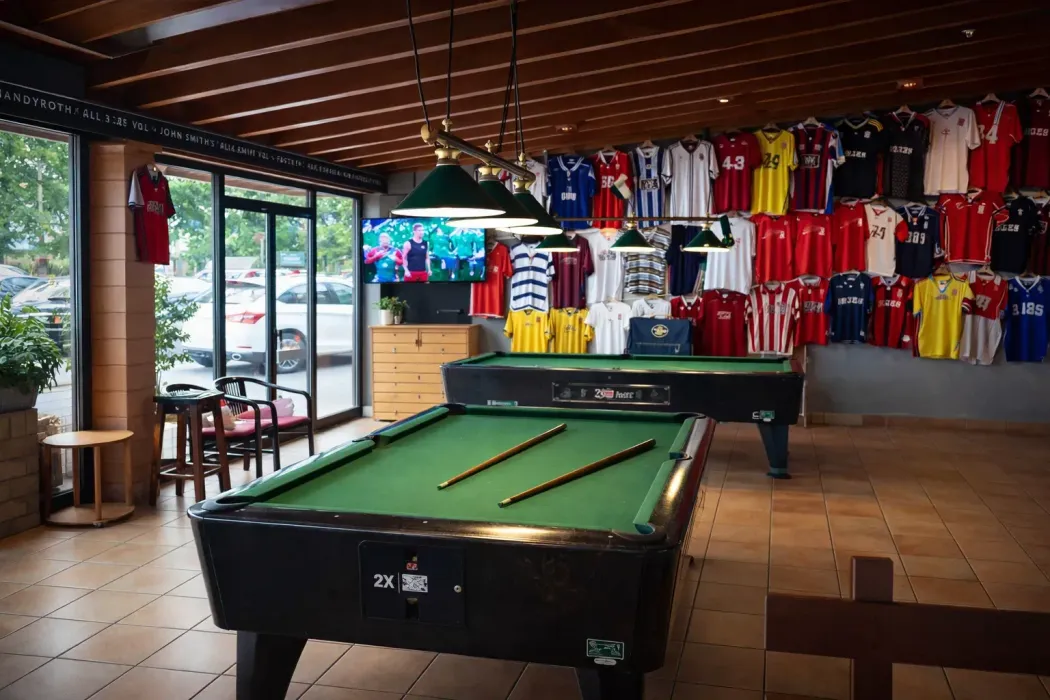 Dos mesas de billar en una habitación decorada con numerosas camisetas deportivas de colores expuestas en la pared.