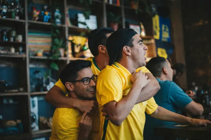 Tres personas con camisetas amarillas celebran mientras ven un partido dentro de un bar con estanterías decoradas al fondo.