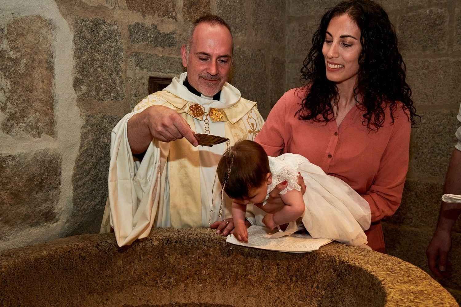 Un hombre con túnica blanca vierte agua sobre un bebé que sostiene sobre una pila bautismal de piedra, junto a una mujer vestida de rosa.