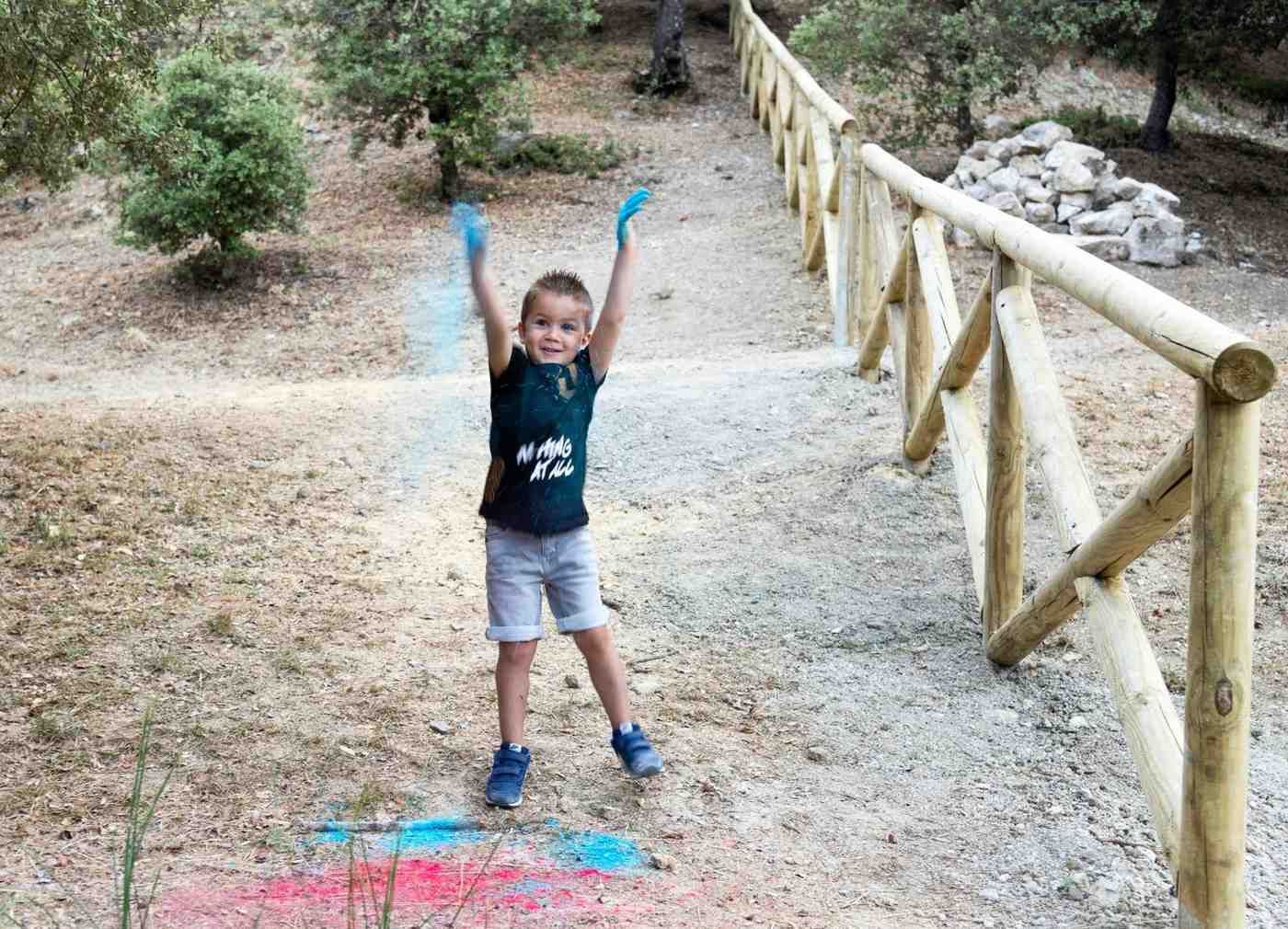 Niño con los brazos levantados en un sendero de tierra junto a una cerca de madera, con polvo azul en el suelo y en las manos.