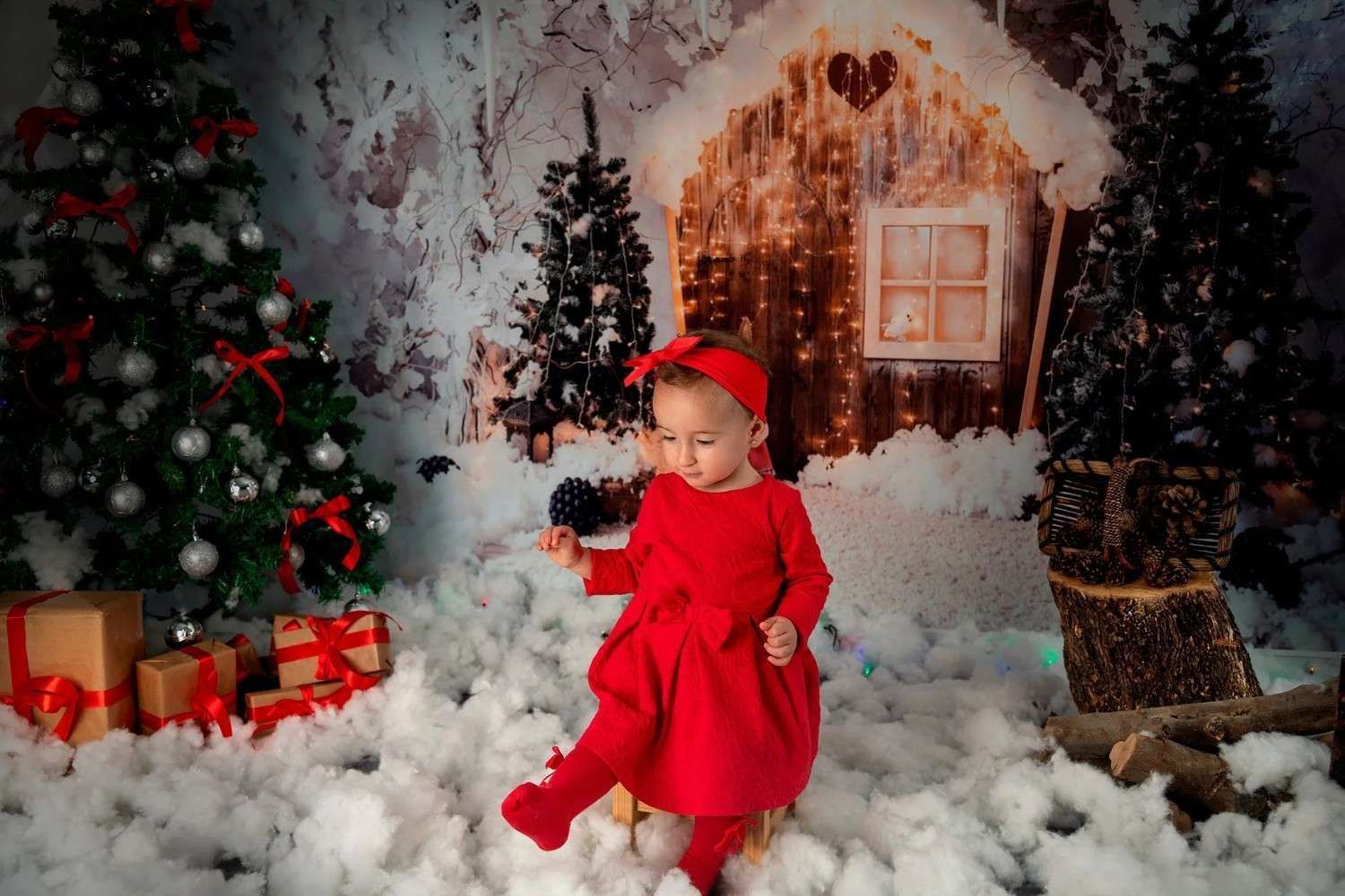 Niña vestida de rojo de pie en una escena navideña nevada con árboles decorados y regalos envueltos.