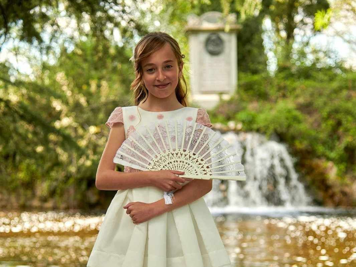 Una niña con un vestido blanco sostiene un abanico decorativo frente a una cascada y una fuente de jardín.