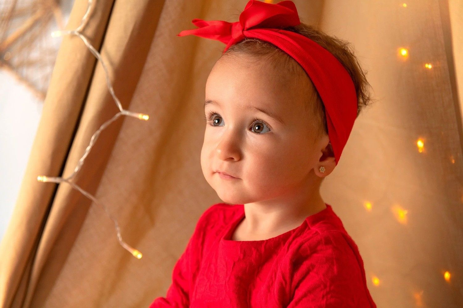 Bebé con diadema y vestido rojos, mirando hacia arriba junto a cálidas luces de hadas y cortinas beige.