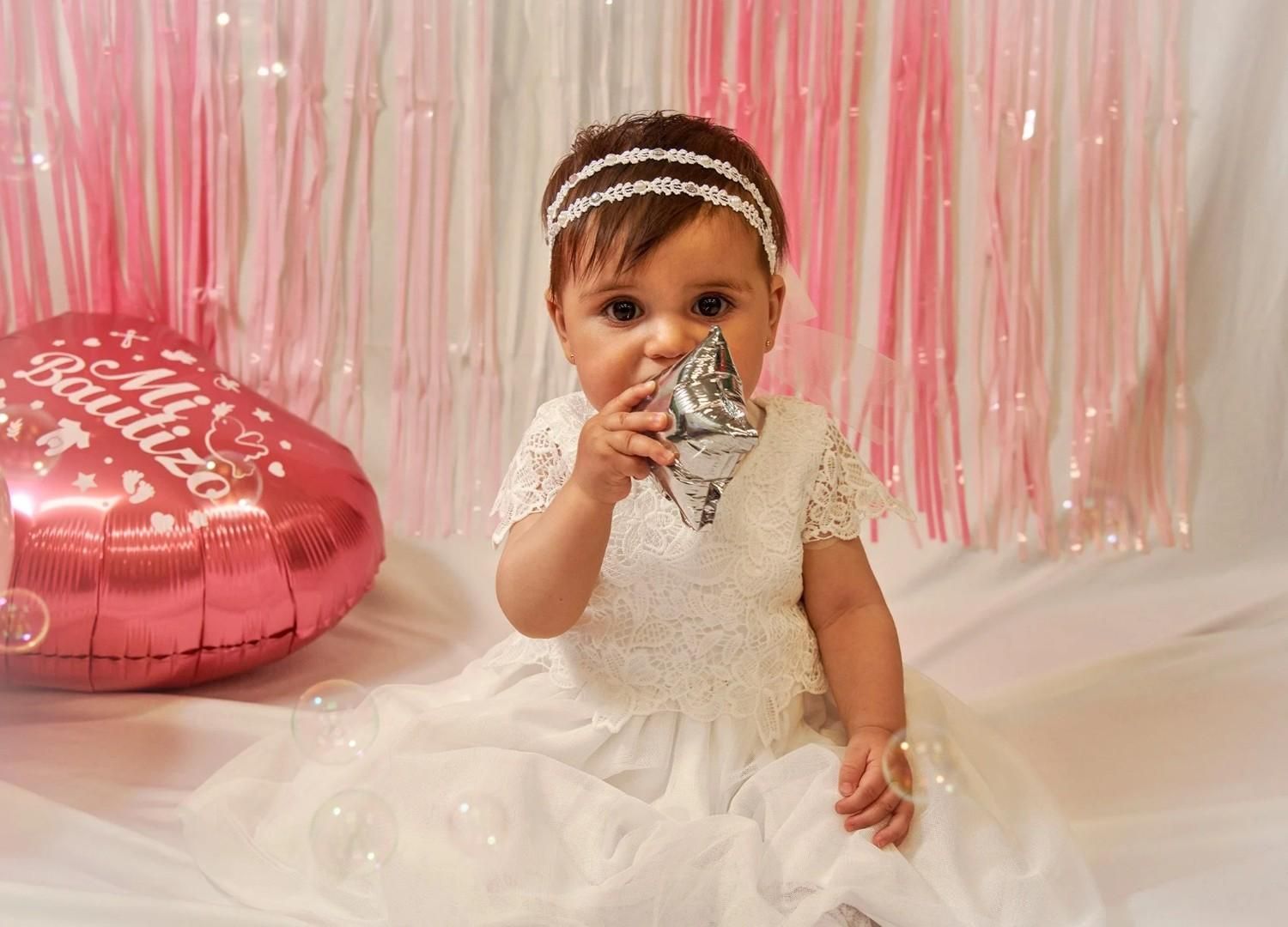 Bebé con vestido blanco sosteniendo un globo plateado, sentada junto a un globo rosa en forma de corazón sobre un fondo rosa.